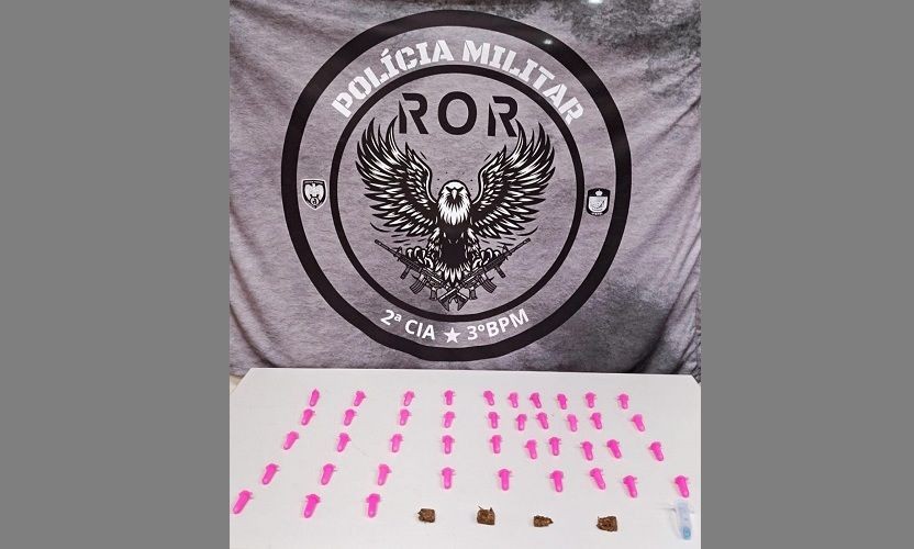 Mesa com pinos de cocaína e buchas de maconha apreendidos pela Polícia Militar de Guaçuí, com ao fundo um banner da corporação durante a operação.