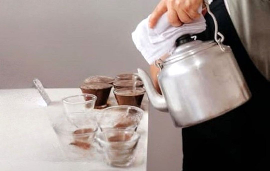 Profissional realiza degustação de café, despejando água quente com chaleira sobre xícaras utilizadas em prova sensorial de cafés especiais.