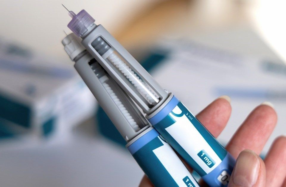 Foto em close-up mostrando uma mão segurando duas canetas aplicadoras de medicamento de cor cinza e azul, com uma pequena agulha visível na ponta superior e um visor digital indicando a dosagem.