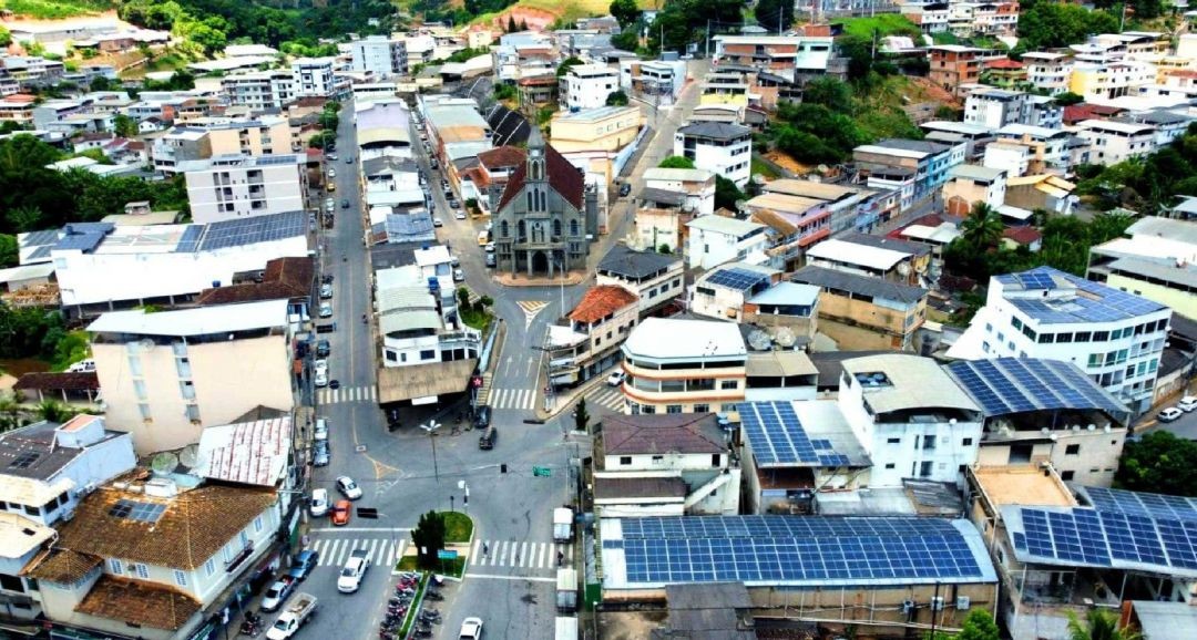 Fotografia aérea do centro de Muniz Freire, destacando a Igreja Matriz ao centro, ruas pavimentadas com faixas de pedestres e diversos prédios comerciais e residenciais, tendo ao fundo, montanhas verdes características do Caparaó.