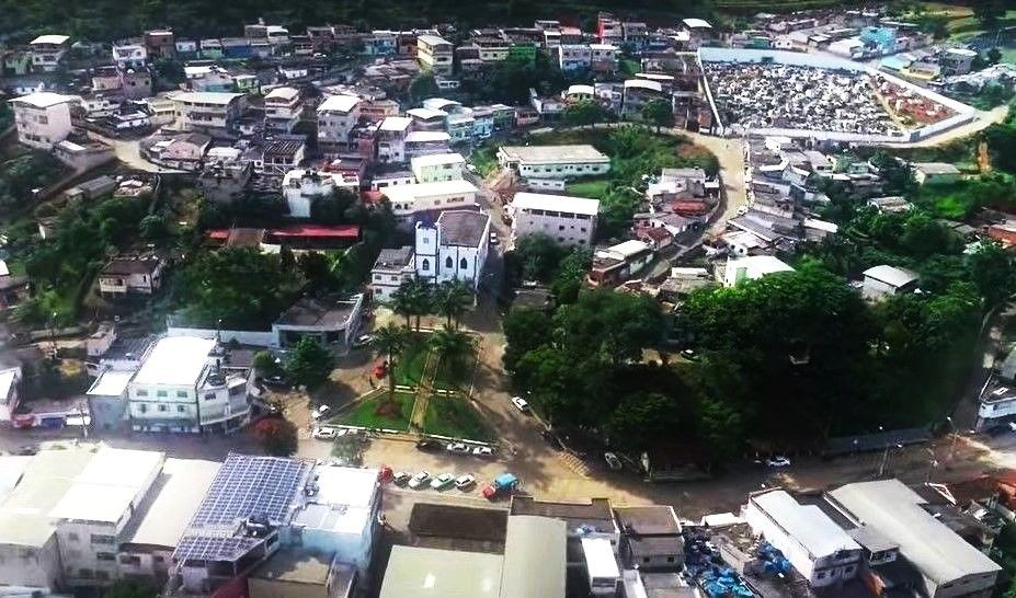 Vista aérea da área central de Dores do Rio Preto, com destaque para a praça, a igreja matriz e prédios do entorno urbano, representando o município reconhecido com o Selo Ouro em alfabetização pelo MEC.