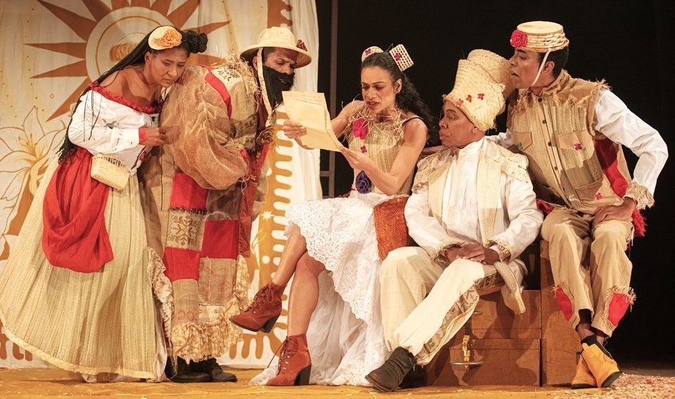 Cinco atores em cena no palco do teatro, vestidos com figurinos rústicos e coloridos, observam atentos enquanto uma personagem sentada lê uma carta, em cenário inspirado na cultura nordestina.