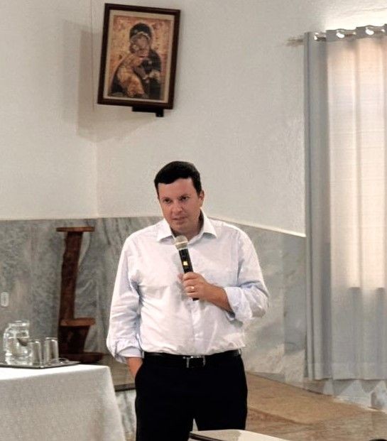 Homem (Promotor Lucas Lobato) de camisa clara e calça escura, segurando um microfone e falando durante a palestra. Ao fundo, uma parede clara com um quadro religioso da Virgem Maria.