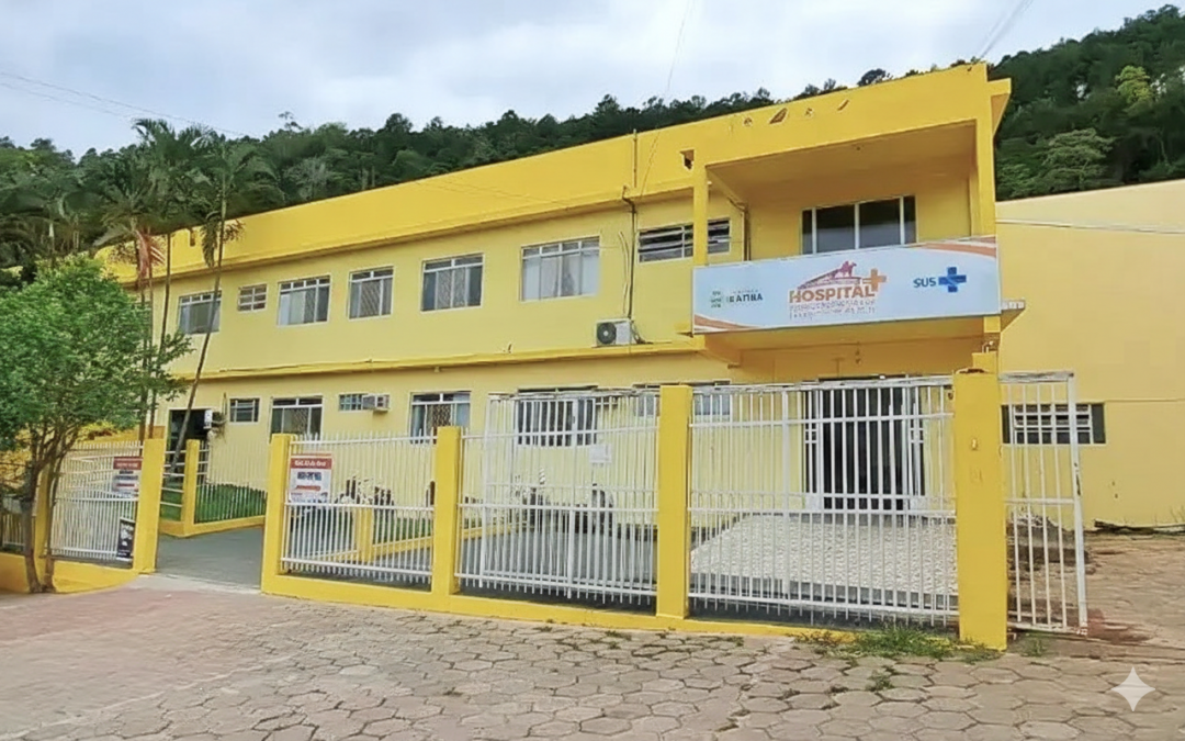 Fachada do Hospital Municipal Dr. Ubirajara Faria Lopes, em Ibatiba, prédio de dois andares pintado de amarelo, com placa de identificação do SUS e grade branca na entrada, localizado em área com vegetação ao fundo.