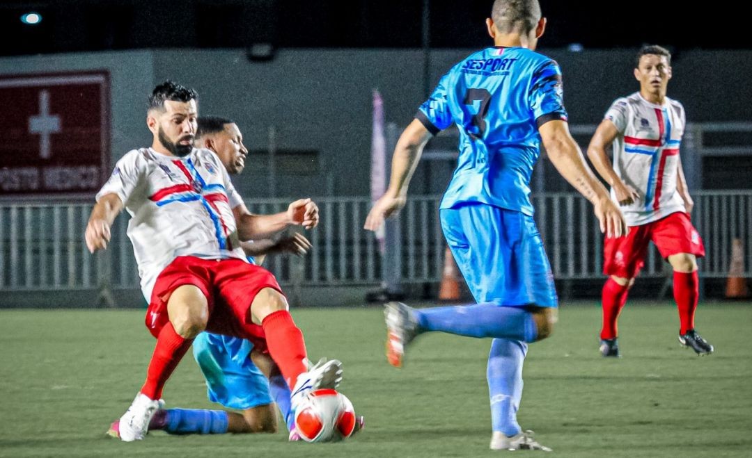 Jogador de uniforme branco e vermelho disputa a bola em lance dividido contra atleta de azul durante partida noturna de futebol amador em campo iluminado.