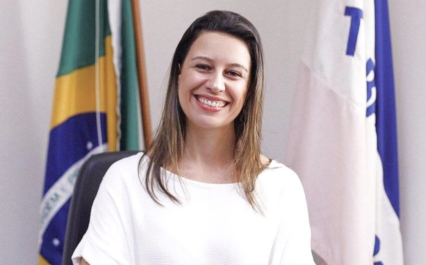 Secretária Cyntia Grillo sorrindo, sentada em gabinete oficial, com bandeiras ao fundo, durante registro institucional.