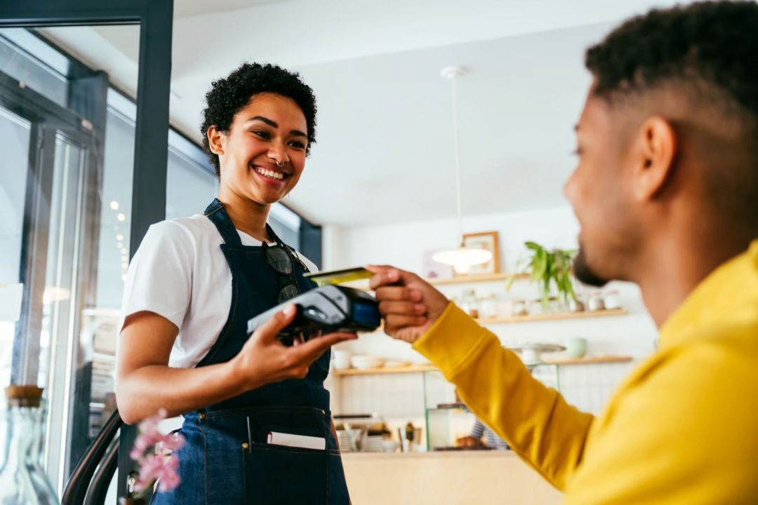 Mulher operadora de caixa realiza pagamento em máquina de cartão enquanto cliente efetua transação, representando consumo e crédito no comércio.