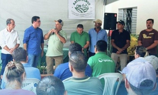 Autoridades municipais, representantes do Projeto Arranjos Produtivos e produtores rurais participam de cerimônia de entrega de mudas em Guaçuí, com agricultor discursando ao microfone e público sentado acompanhando o evento.