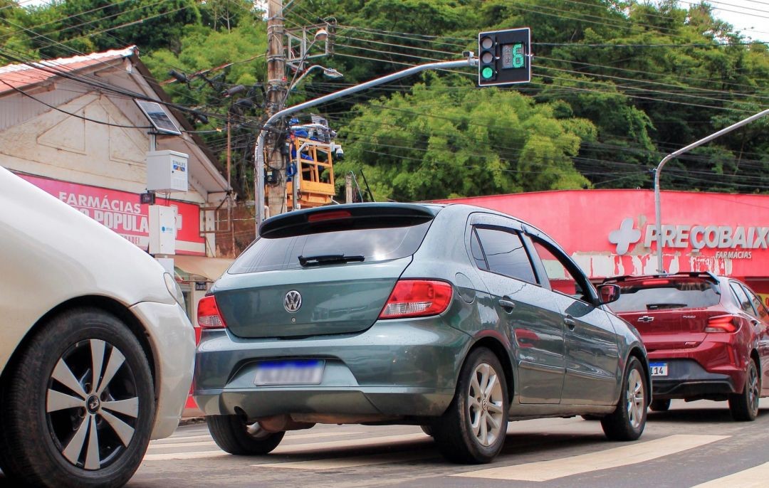 Carro Volkswagen Gol cinza parado na faixa de pedestres em frente a uma farmácia, aguardando o semáforo que exibe o cronômetro digital na cor verde.