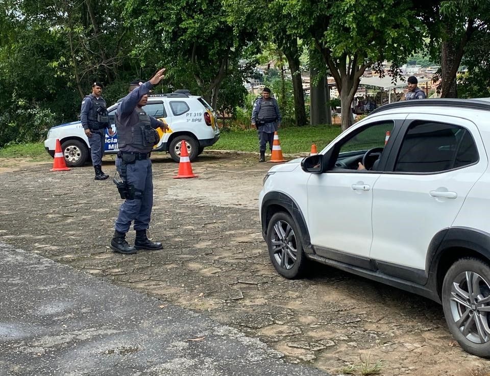 Policial militar realiza abordagem durante blitz de trânsito com cones de sinalização e viatura ao fundo.