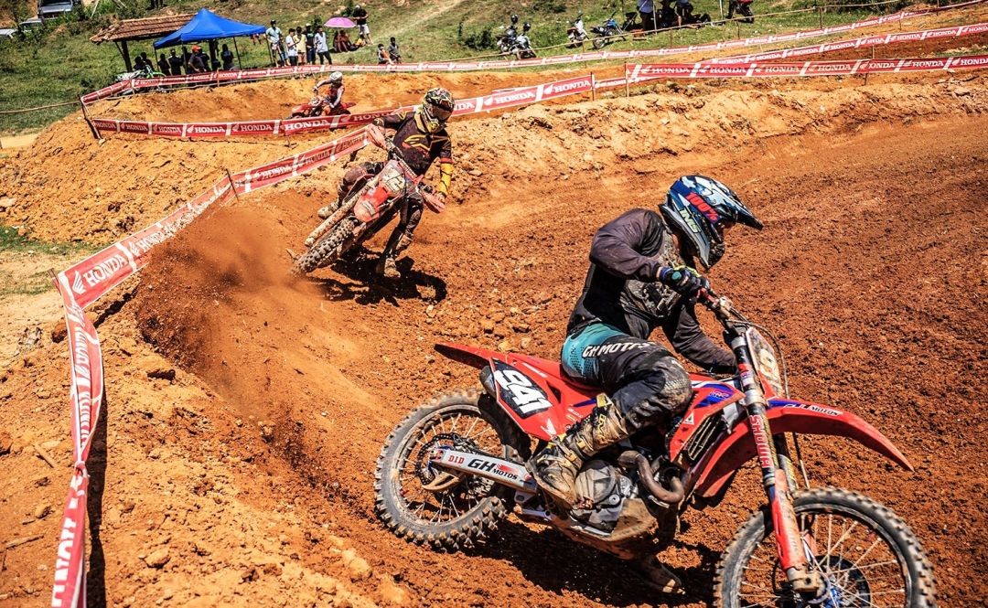 Vista lateral de dois pilotos de motocross em uma curva acentuada de terra laranja. O piloto em primeiro plano usa uniforme azul e preto, em uma moto vermelha com o número 941. Ao fundo, outro piloto em uma moto Honda levanta poeira na curva. A pista é cercada por fitas de isolamento da marca Honda e há público ao fundo sob tendas.