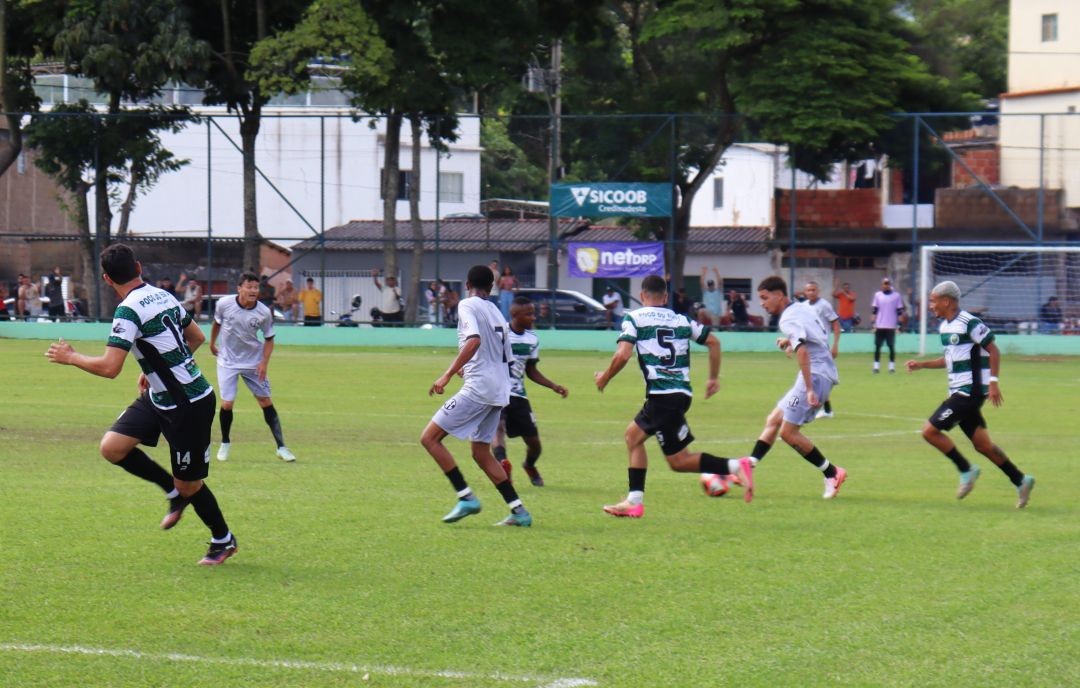 Jogadores de futebol em campo durante partida do Campeonato Sicoob Credisudeste em Dores do Rio Preto. Atletas vestem uniformes listrados em verde e branco e uniformes cinzas; ao fundo, arquibancada com público e banners dos patrocinadores.