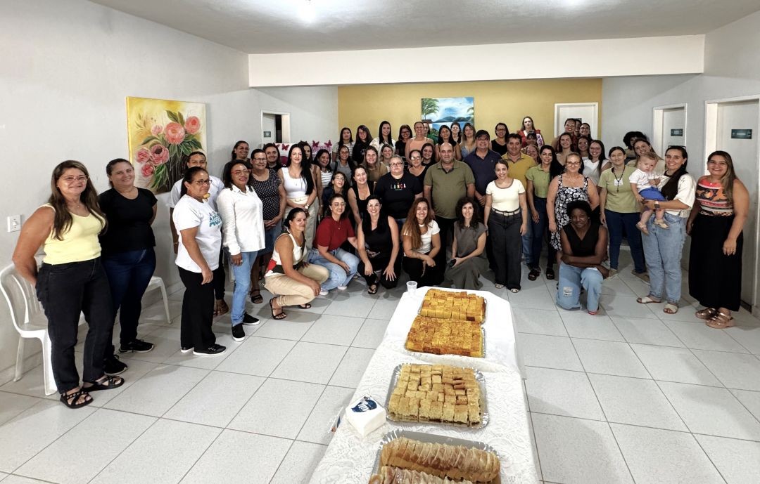 Grande grupo de mulheres servidoras da Prefeitura de Divino de São Lourenço posando para foto oficial em um salão. À frente, uma mesa com pães e bolos para o café da manhã. Ao centro, o prefeito Dudu Queiroz e autoridades locais.
