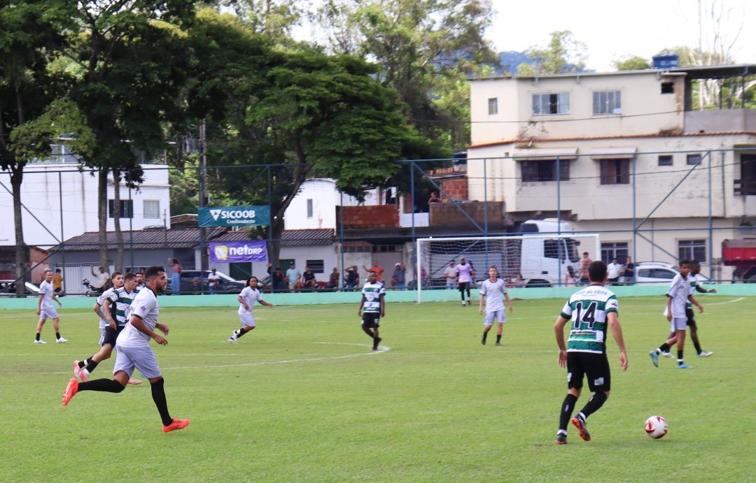 Partida do Campeonato Sicoob Credisudeste de Futebol de Campo em andamento, com jogadores disputando a bola em campo gramado cercado por arquibancada e casas ao fundo.