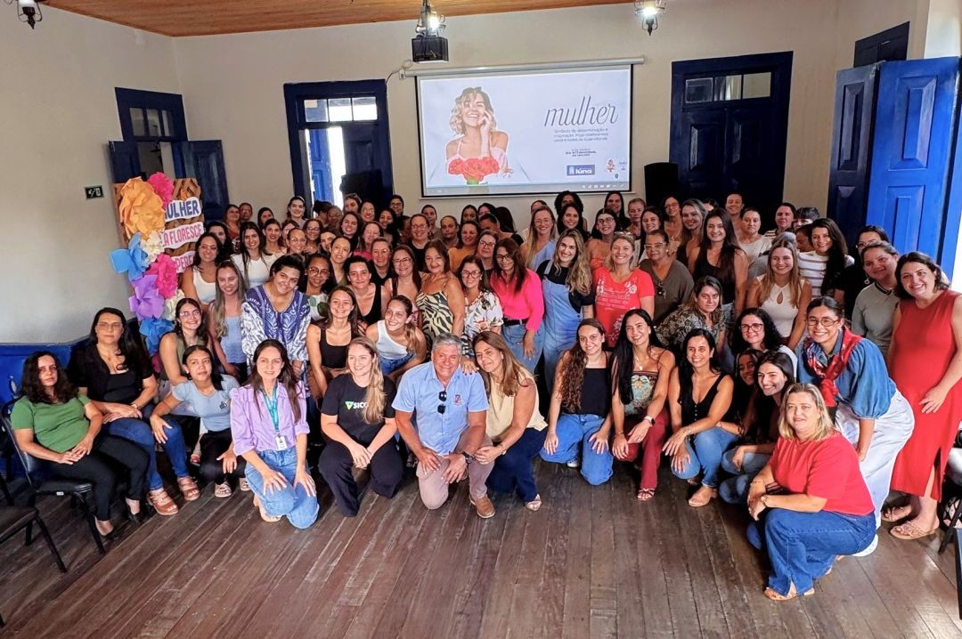 Fotografia em grupo no interior da Casa da Cultura de Iúna. Dezenas de mulheres, servidoras municipais, posam sorridentes junto ao prefeito Romário Vieira, que está agachado ao centro. Ao fundo, um telão exibe uma arte de homenagem ao Dia da Mulher e há um painel decorativo com flores coloridas de papel.