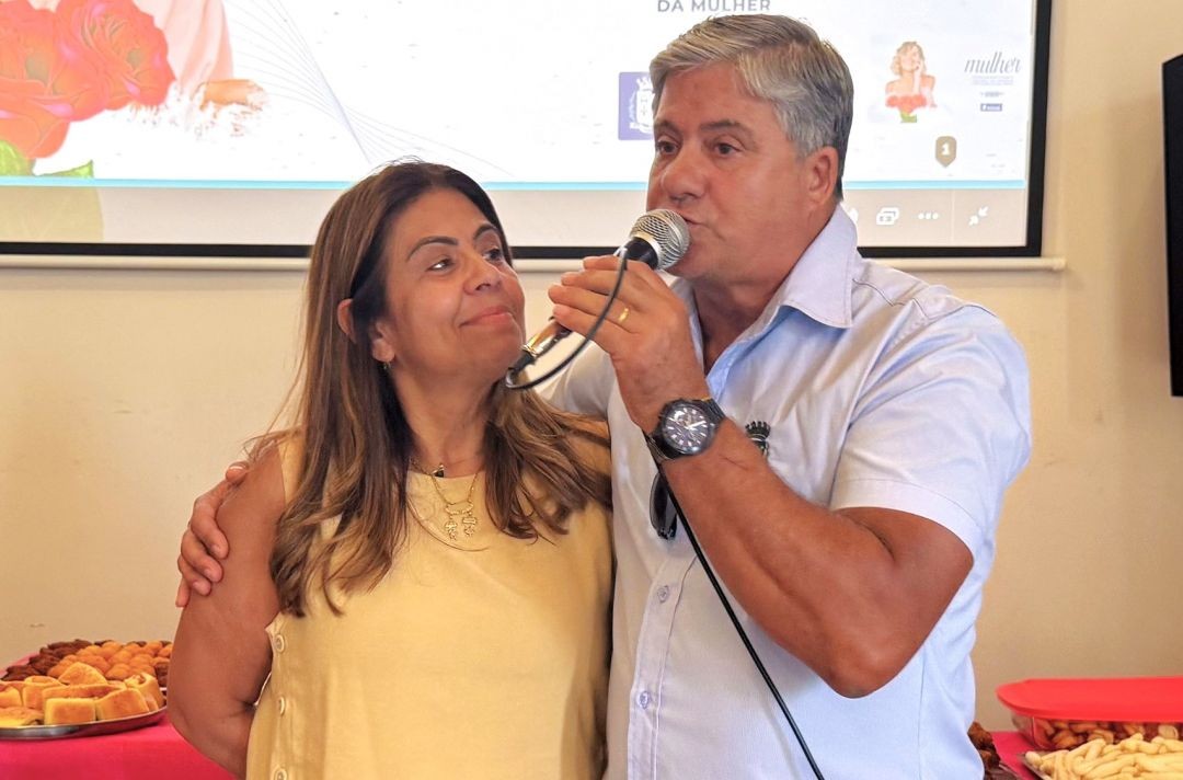 Close do prefeito Romário Vieira falando ao microfone durante o evento. Ele está com o braço em volta dos ombros da primeira-dama e secretária de Assistência Social, Lusmar Vieira (Tuca), que o olha com um sorriso. Ao fundo, mesas com café da tarde e o telão do evento.