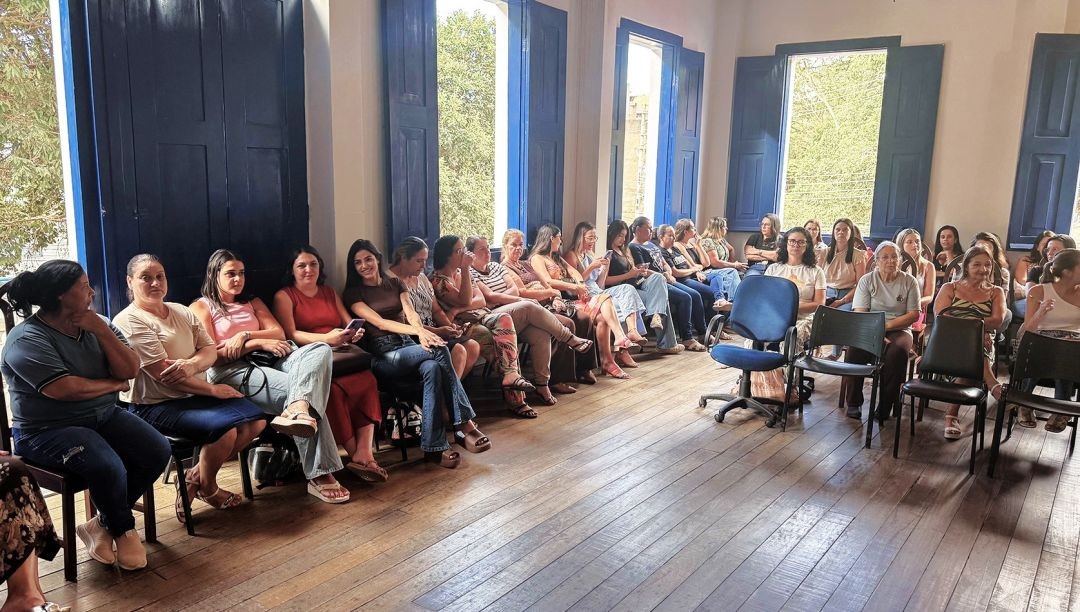 Fotografia em plano médio capturando uma fileira de servidoras municipais sentadas em cadeiras ao longo da parede esquerda de uma sala ampla na Casa da Cultura. O chão é de tacos de madeira e ao fundo veem-se janelas altas com portais azuis. As mulheres estão vestidas com roupas casuais e olham atentamente para a direita da imagem, indicando que acompanham o início de uma apresentação.
