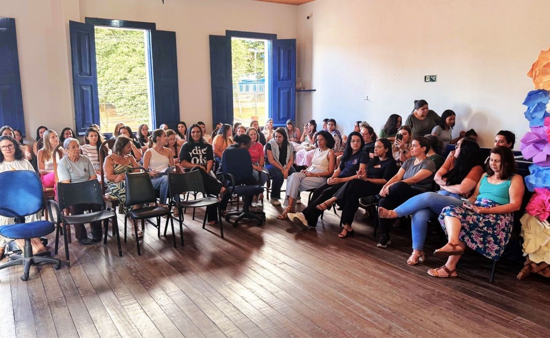 Fotografia em plano geral mostrando a lateral direita de uma sala de conferência na Casa da Cultura de Iúna. Diversas servidoras municipais estão sentadas em cadeiras dispostas em filas, participando de um evento. O ambiente possui um piso de madeira rústica e janelas com molduras e portas azuis ao fundo, revelando a claridade do dia e árvores do lado de fora. Na parede da direita, próximo às servidoras, há uma decoração vertical colorida feita de flores de papel em tons de laranja, azul e rosa.
