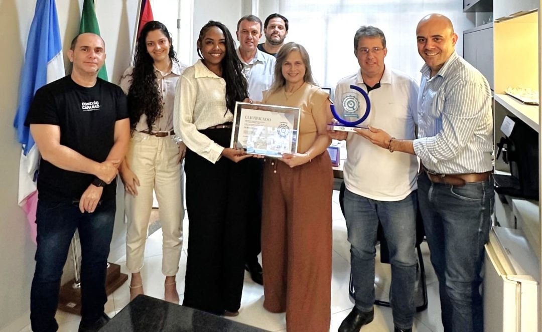 Grupo de sete pessoas em um escritório, posando para foto. Ao centro, duas mulheres e um homem seguram um certificado emoldurado e um troféu azul em formato de semicírculo (Selo Diamante). À esquerda, bandeiras oficiais. Todos sorriem, vestindo roupas casuais e profissionais.