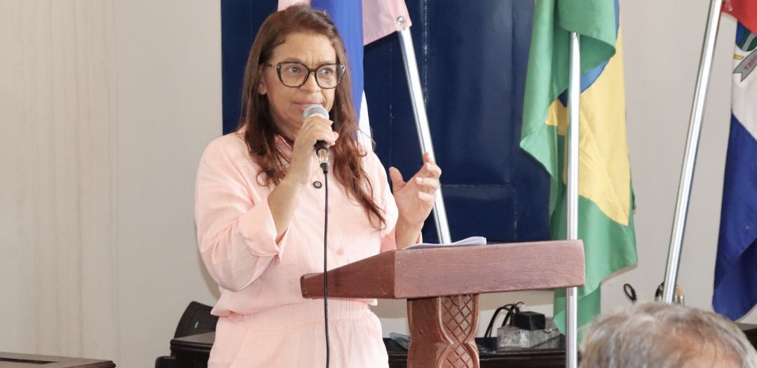 A Secretária Municipal de Controle e Transparência, Elzeni da Silva Oliveira, discursando na solenidade de entrega do Selo Diamante de Transparência, em púlpito de madeira e, ao fundo, bandeiras do Espírito Santo, Brasil e Iúna.