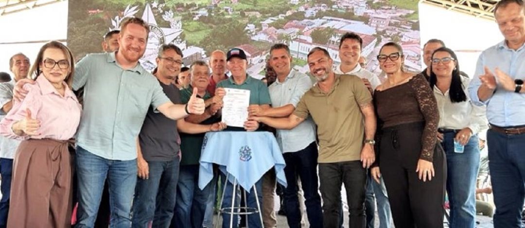 Fotografia colorida de ângulo aberto mostrando um grande grupo de autoridades, incluindo o governador Renato Casagrande e o prefeito de São José do Calçado, posicionados sob uma tenda de eventos. Eles seguram um documento assinado sobre uma mesa forrada com uma toalha azul com o brasão estadual. Ao fundo, um painel exibe uma foto aérea do município.