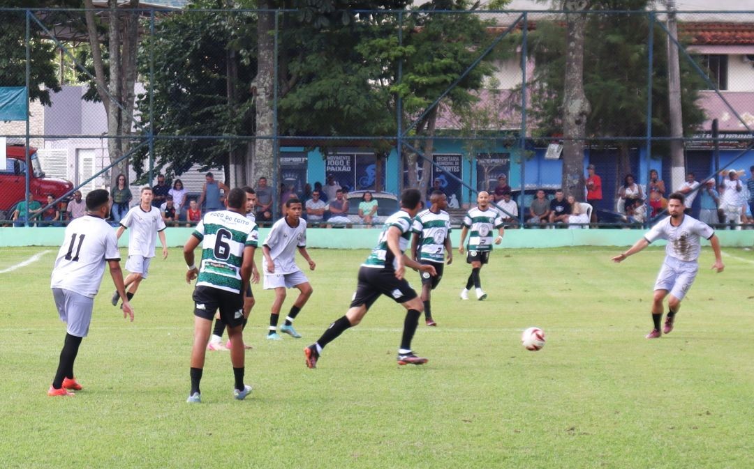 Fotografia de uma partida de futebol de campo durante o dia. Jogadores vestindo uniformes em tons de branco, cinza e listras verdes e pretas disputam a posse da bola no centro do gramado verde. Ao fundo, vê-se uma tela de proteção, árvores e o público acompanhando o jogo sentados em bancos próximos a uma construção azul com a frase 