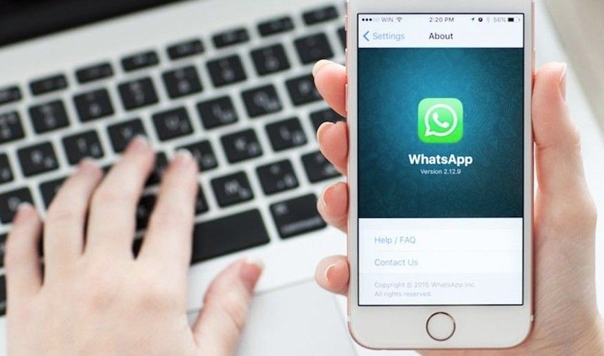 Fotografia em close de uma pessoa segurando um iPhone branco que exibe o logotipo do WhatsApp na tela. Ao fundo, fora de foco, as mãos de outra pessoa digitam em um teclado de notebook prata.