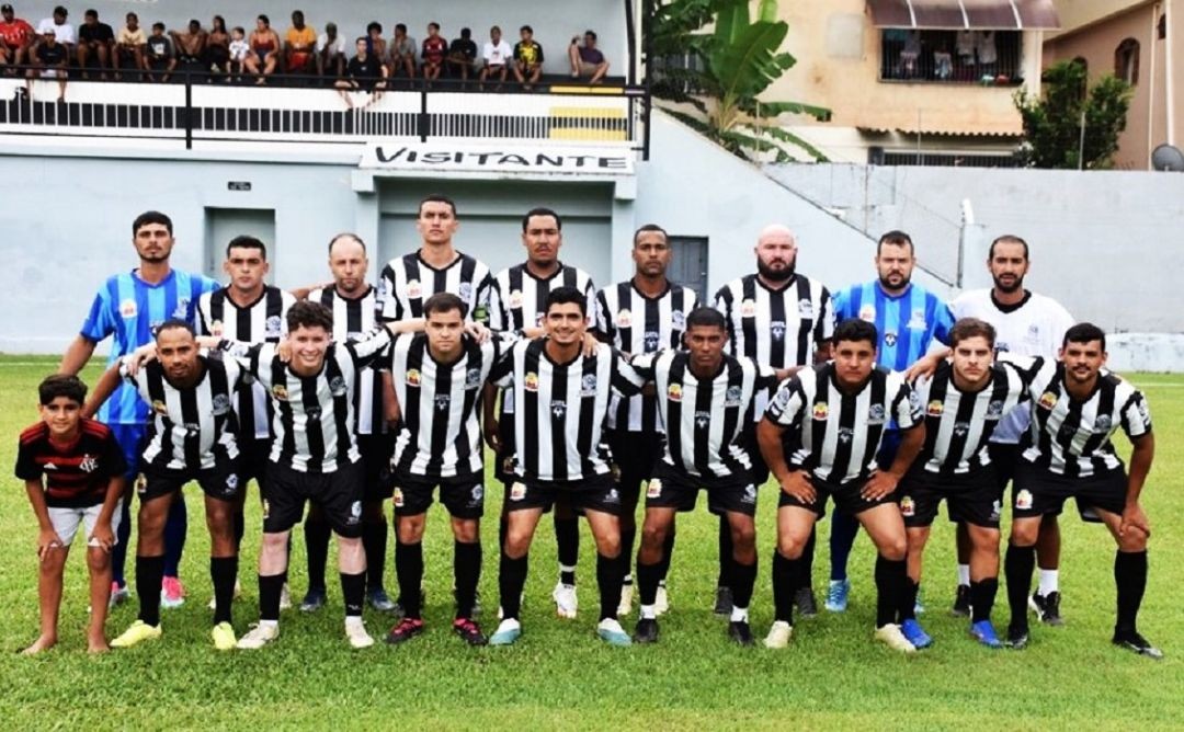 Registro da equipe masculina de Irupi no gramado. O time veste uniforme com listras verticais pretas e brancas (estilo tradicional). Jogadores aparecem abraçados e perfilados. Ao fundo, é possível ver uma arquibancada com torcedores e uma placa escrita 