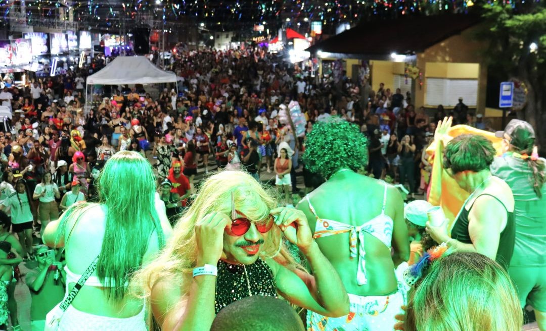 Fotografia em plano médio elevada mostrando uma multidão em uma rua enfeitada à noite. No primeiro plano, pessoas fantasiadas em cima de um trio elétrico ou palco, com destaque para uma pessoa de peruca loira, óculos escuros vermelhos e maquiagem, sob luzes verdes. Ao fundo, o público ocupa toda a extensão da via sob cordões de luzes coloridas.