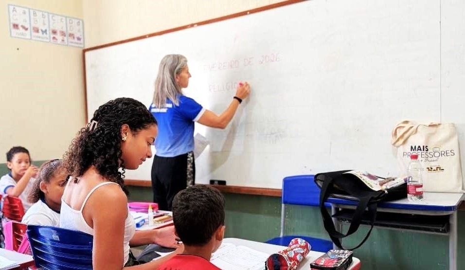 Mulher loira com camisa azul de uniforme escreve no quadro branco de uma sala de aula. À frente, em primeiro plano, uma jovem de cabelos cacheados e uma criança observam a atividade. No quadro, lê-se 