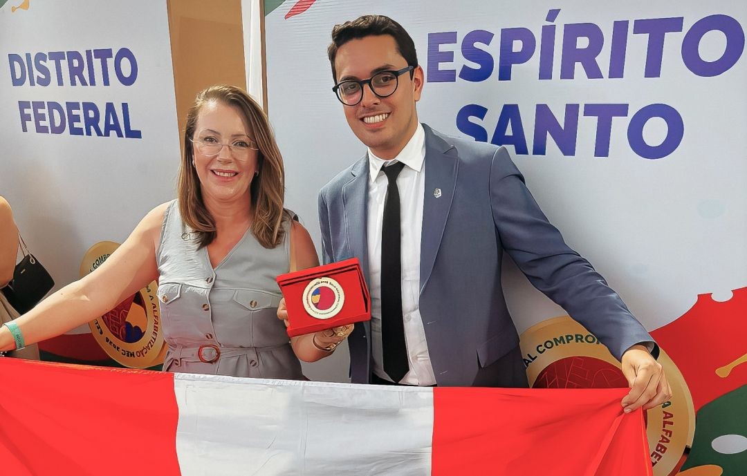 Vice-prefeito e secretária de Educação de Irupi exibem medalha do Selo Ouro do MEC em Brasília, segurando a bandeira do município durante cerimônia do programa Criança Alfabetizada.