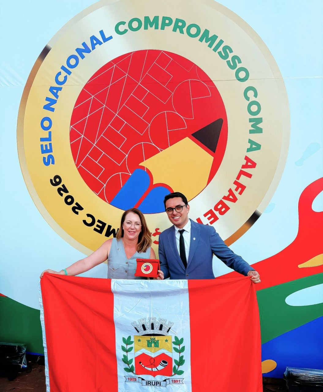 Secretária de Educação e vice-prefeito de exibem medalha do Selo Ouro do MEC em Brasília, segurando a bandeira do município, tendo ao fundo um painel com a logomarca do programa “Selo Nacional Compromisso com a Alfabetização – MEC 2026.