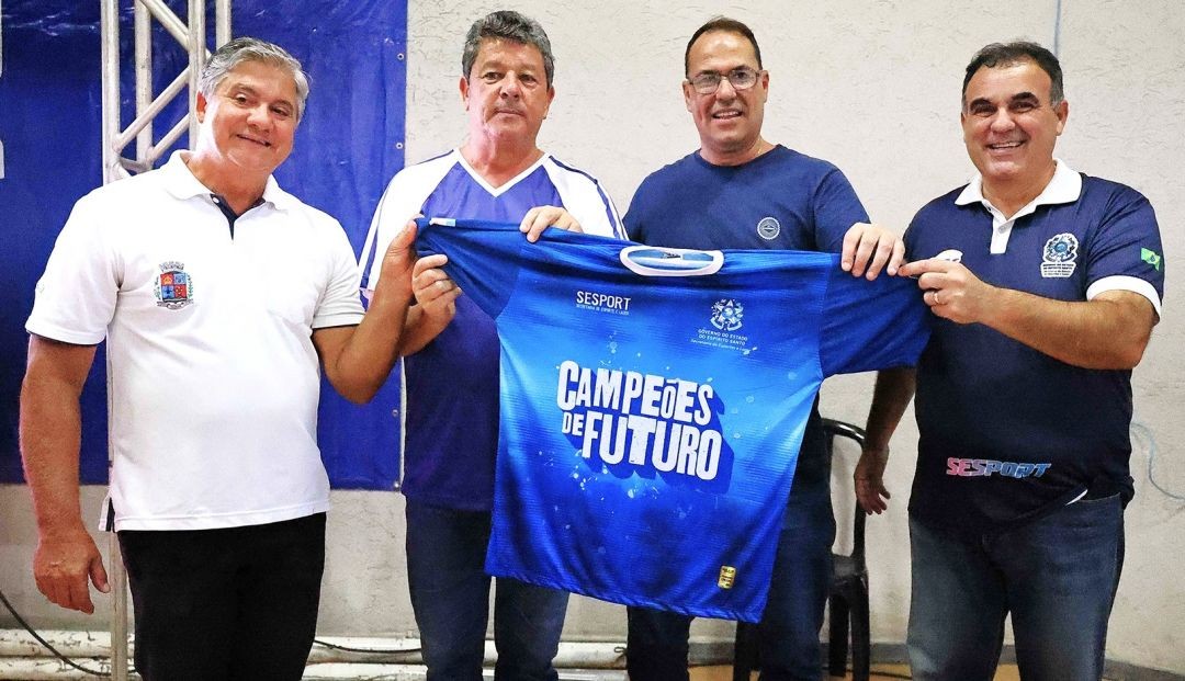 O prefeito Romário Viera, o secretário Zizinho, vereador Adilsinho e André Varão posando para, segurando uma camisa azul com as palavras “Campeões do Futuro”, em destaque e, também, “Sesport” e o brasão do Governo do Estado do Espírito Santo.