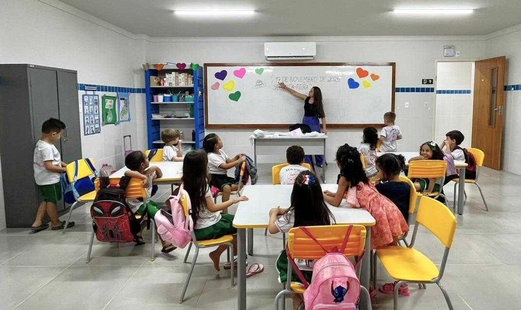 Sala de aula com professora à frente de um quadro branco e alunos sentados em mesas, em ambiente escolar de educação infantil.