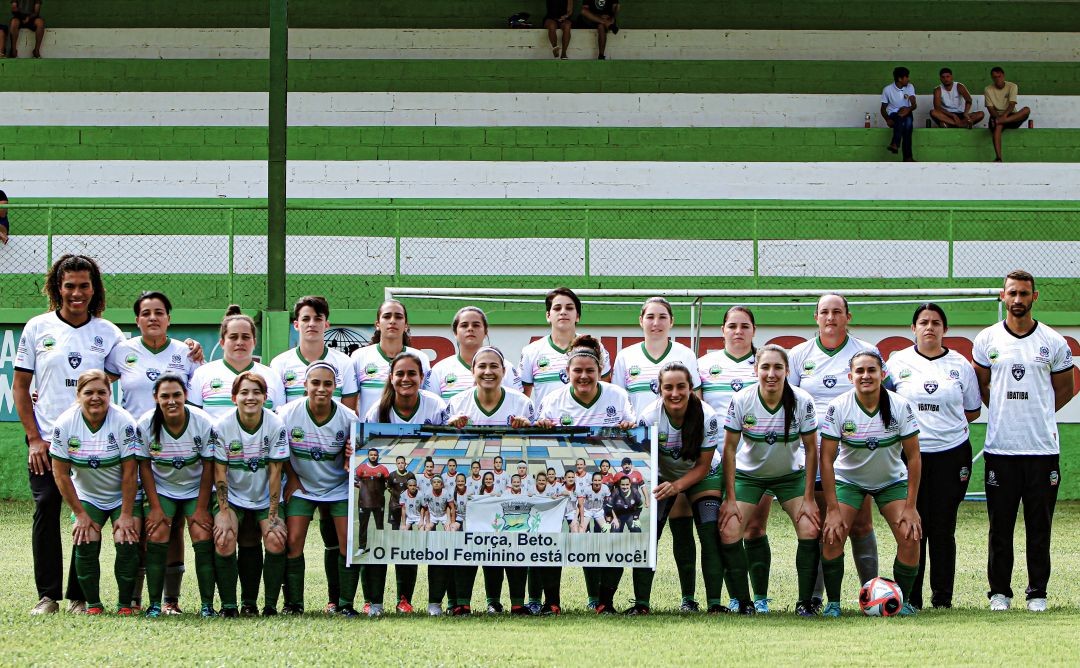 Equipe de futebol feminino de Ibatiba posa alinhada no campo antes da partida, segurando faixa de apoio, com arquibancada ao fundo.