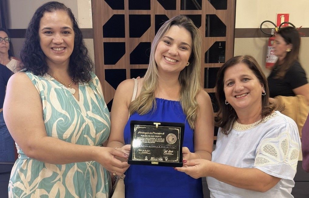 A secretária Esther Simões entre Elizabeth Gomes e Ana Claudia Escolarique. Elas sorriem para a foto em um ambiente interno e Esther, vestindo uma blusa azul, segura uma placa de homenagem escura com detalhes dourados. À esquerda, Elizabeth veste blusa estampada em tons de verde e branco, e à direita, Ana Claudia veste blusa branca com detalhes rendados.
