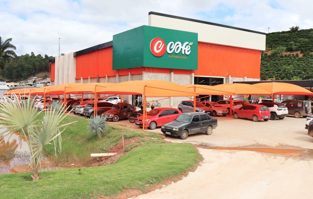 Vista externa do Café Supermercados em Irupi. O prédio possui cores vibrantes em laranja e verde, com um grande painel com a logo da empresa. À frente, um amplo estacionamento coberto com toldos laranjas está repleto de carros. O ambiente é cercado por gramado verde e palmeiras.