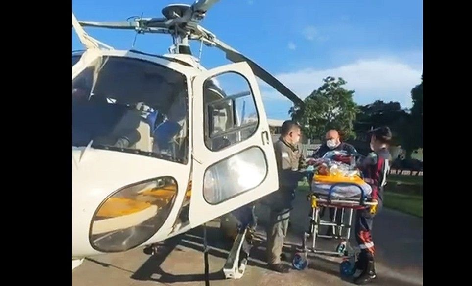 Helicóptero do NOTAER com porta aberta durante atendimento aeromédico, enquanto equipe realiza transferência de criança em maca ao lado de profissionais de resgate em área de pouso.