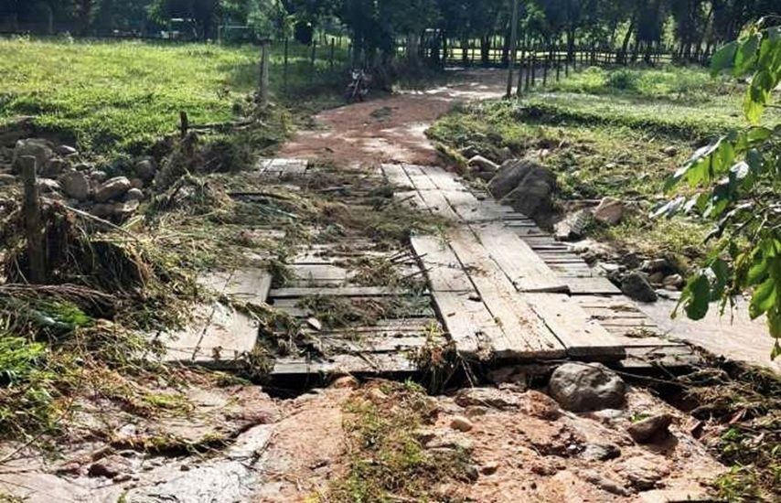 Ponte de madeira em área rural danificada pela enxurrada, com acúmulo de galhos, lama e pedras sobre a estrutura e nas margens.