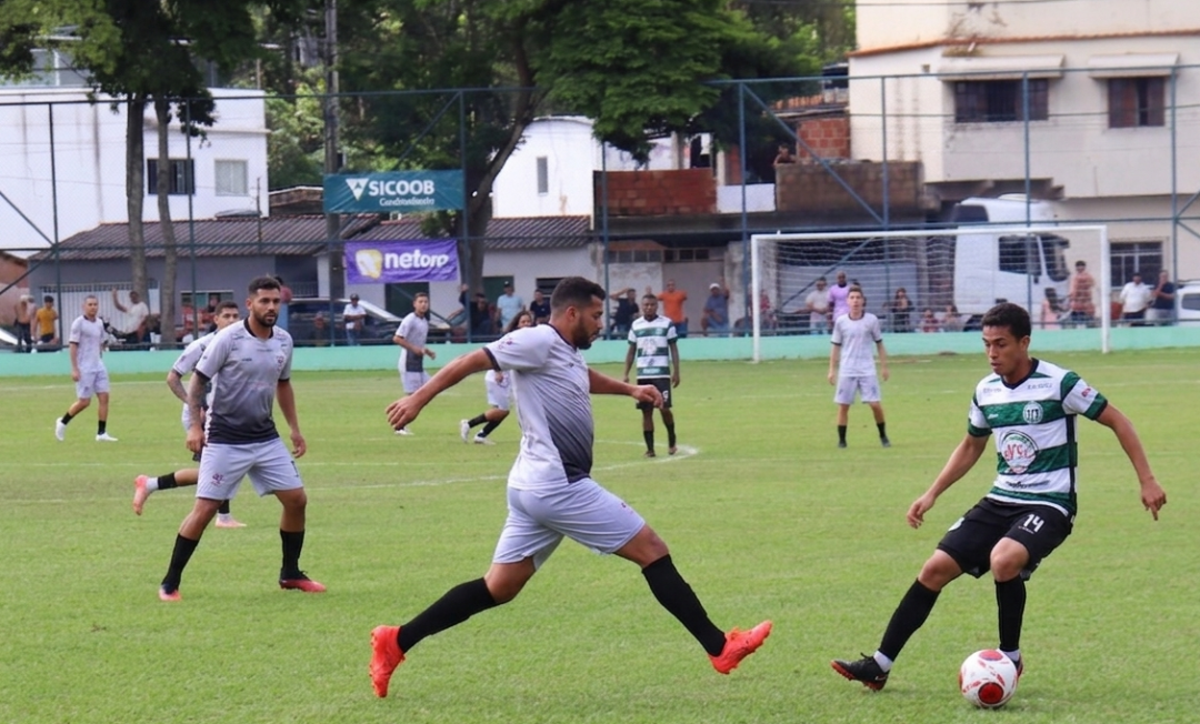 Jogadores disputam bola em partida de futebol amador no Estádio Municipal de Dores do Rio Preto, com equipes uniformizadas e público ao fundo.