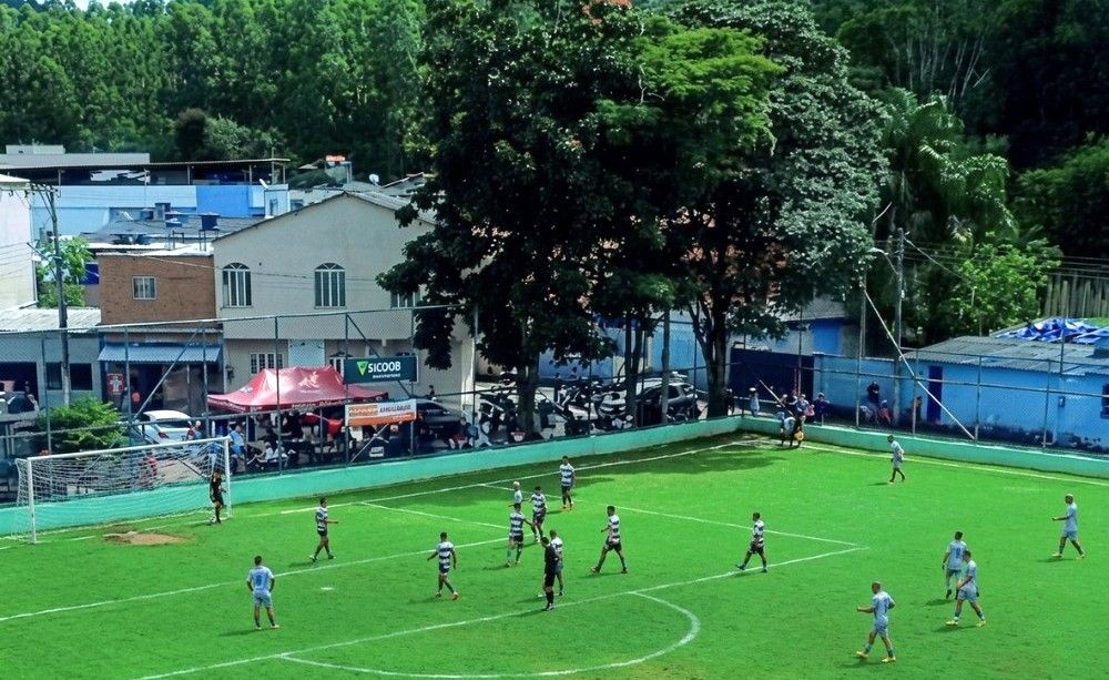 Uma fotografia de ângulo elevado em alta resolução captura uma partida de futebol de campo no Estádio Municipal de Dores do Rio Preto. O campo, de grama verde vibrante e linhas brancas nítidas, está preenchido por jogadores vestidos com uniformes azuis e cinzas claros, claramente definidos e em movimento. O gol está visível à esquerda. Ao fundo, uma área urbana com edifícios detalhados, uma barraca de fornecedor vermelha com texto legível e grandes árvores frondosas que cobrem uma encosta florestal.