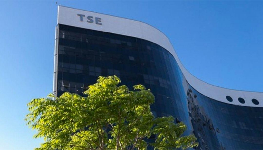Vista parcial da fachada do prédio do Tribunal Superior Eleitoral (TSE), em Brasília, com estrutura moderna e árvore verde no centro da imagem, com céu azul ao fundo.
