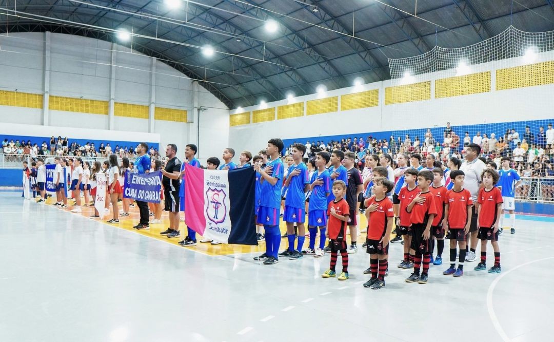 Vista ampla do ginásio com diversas delegações de estudantes perfiladas na quadra. À frente, alunos da Escola Santíssima Trindade seguram uma bandeira rosa, azul e preta. Os jovens vestem uniformes esportivos de diferentes cores e estão com a mão no peito para o hino.