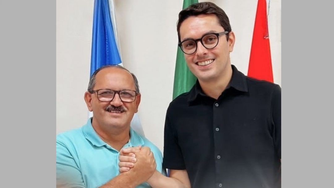 Prefeito Paulino Lourenço e vice-prefeito Abercílio Machado, de Irupi, aparecem lado a lado em ambiente institucional, sorrindo e cumprimentando-se com um aperto de mãos, com bandeiras ao fundo.