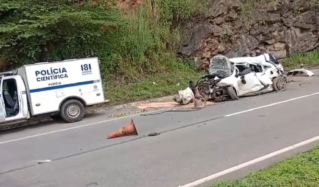 Imagem lateral de carro branco destruído após colisão contra uma ribanceira na Rodovia ES-482, com presença de viatura da Polícia Científica e um cone de sinalização derrubado na pista.