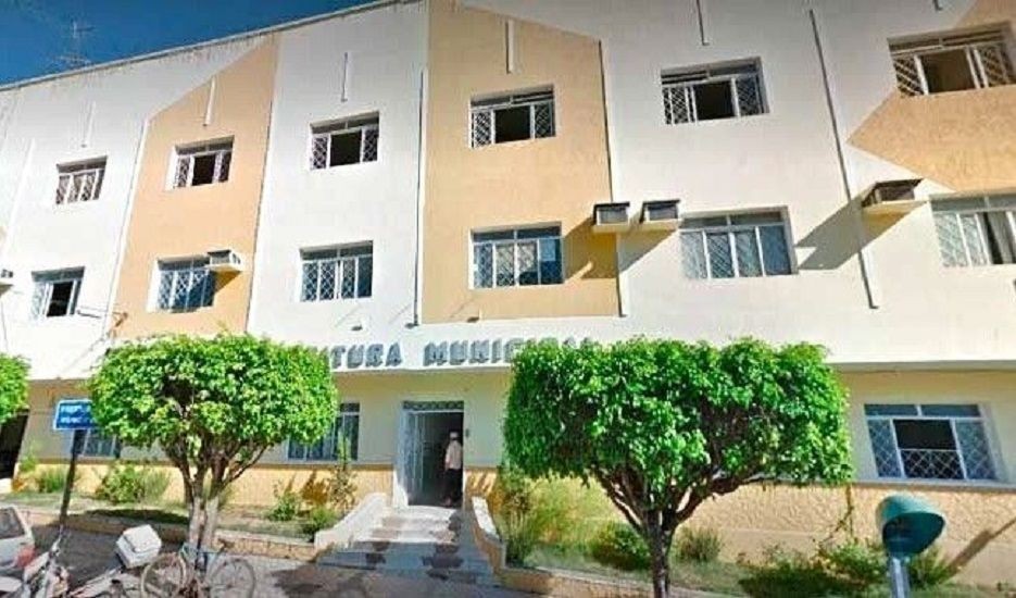 A imagem mostra a fachada da Prefeitura Municipal de Iúna. O prédio é branco com detalhes em tom bege/ocre, possui três pavimentos e janelas com grades brancas. À frente, há duas árvores de copa redonda e verde, uma bicicleta estacionada à esquerda e um orelhão (telefone público) azul à direita. A placa 