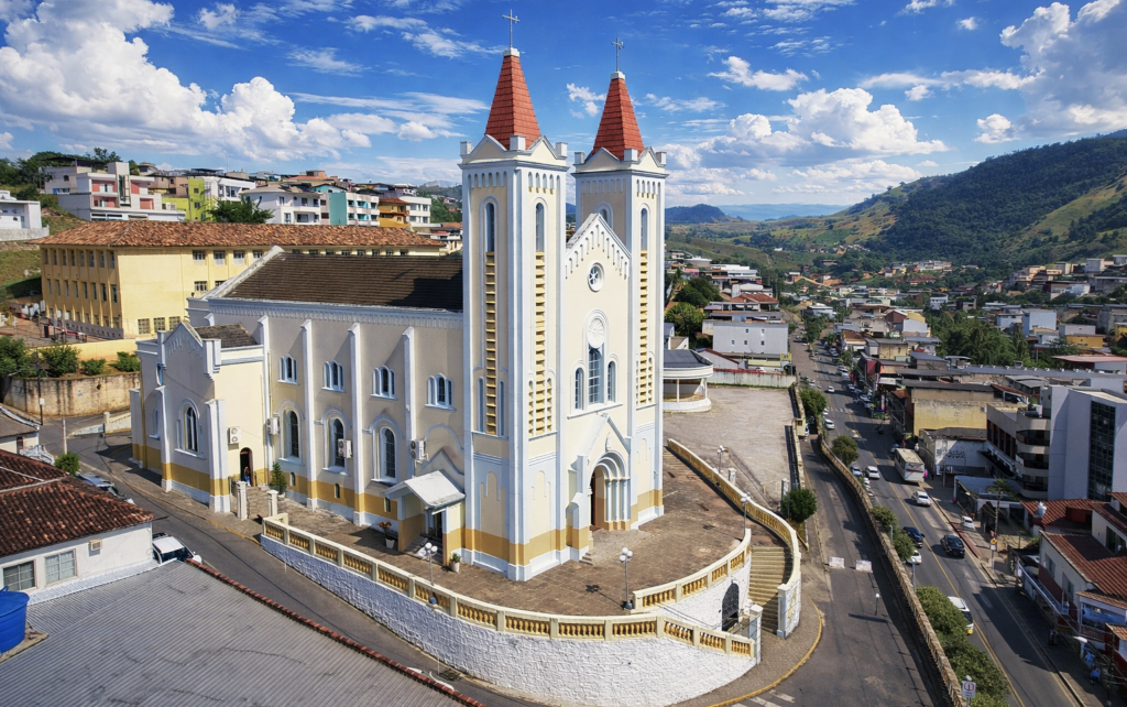 Vista aérea atual da Paróquia Nossa Senhora da Penha em Alegre. O templo possui cor bege com detalhes em branco e telhados das torres em tom alaranjado. Ao fundo, observa-se a paisagem urbana de Alegre e as montanhas verdes do Caparaó.