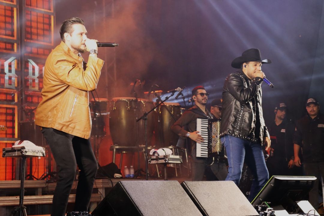 Dois cantores se apresentam no palco durante show da Expoagro de Patrimônio da Penha, acompanhados por músicos e instrumentos ao fundo, com iluminação cênica e público fora do enquadramento.
