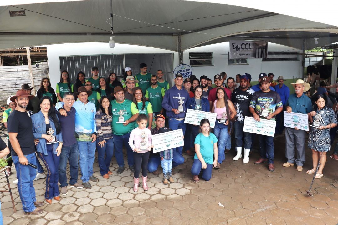 Grupo de cerca de 30 pessoas, entre homens, mulheres e crianças, posam para foto oficial sob uma tenda branca. Alguns seguram troféus dourados e cheques simbólicos de premiação. Ao fundo, vê-se a estrutura de um pavilhão rural.