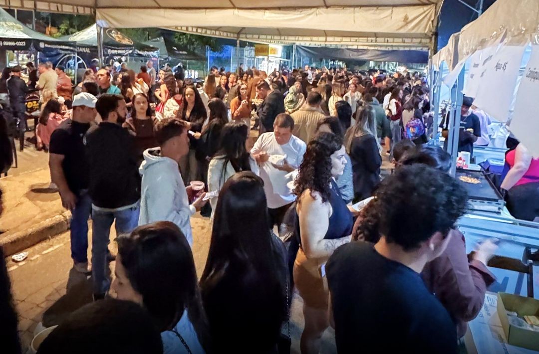 Fotografia noturna da Feira Arte e Sabor em Iúna. Diversas pessoas circulam por uma rua coberta por tendas brancas, onde funcionam barracas de comida e bebidas. O ambiente está movimentado e iluminado.